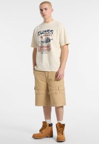 T-shirt grafico beige con design stampato, abbinato a pantaloni cargo color cammello con molteplici tasche. Indossato con stivali marroni con lacci.