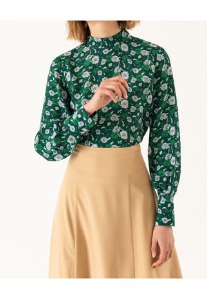 Personne portant un chemisier vert à fleurs à manches longues avec col montant, associé à une jupe beige taille haute sur un fond uni.