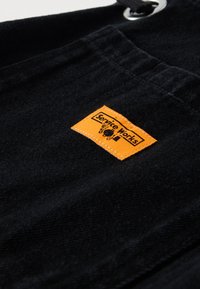 Czarna tkanina o fakturowanej powierzchni z wyraźną pomarańczową metką "Service Works" z czarnym tekstem i grafiką.