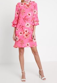 Nainen, joka pukeutuu vaaleanpunaiseen kukalliseen polvipituiseen mekkoon, jossa on röyhelösateet, ja valkoisiin korkokenkiin, seisoo yksinkertaista taustaa vasten.