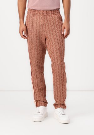 Homme portant un pantalon marron à motif géométrique avec des bandes latérales, associé à des baskets blanches et une chemise rose clair.