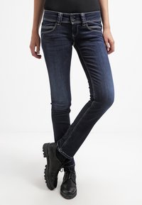 Kvinna som bär mörkblå slim-fit jeans och svarta snörskor, stående med benen korsade mot en vit bakgrund.