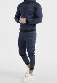 Marineblauwe trainingspak gemaakt van rekbare stof met een rits hoodie en nauwsluitende joggingsbroek, voorzien van ribgebreide manchetten en merkaccenten.