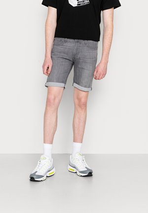 Shorts de mezclilla gris con dobladillos enrollados, combinados con una camiseta gráfica negra y zapatillas gris-blancas con acentos neón. Diseño sólido y casual.