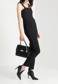 Svart ärmlös jumpsuit med åtsittande överdel och vida ben, ihopparad med en svart handväska med nitar och spetsiga svarta ankelstövlar.