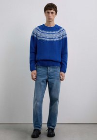 Suéter azul de punto con patrones geométricos blancos a lo largo del pecho, combinado con jeans azul claro y zapatos negros. Diseño sencillo y casual.