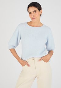 Maglione a coste blu chiaro, con maniche corte e a sbuffo. Texture morbida e scollatura rotonda. Indossato con pantaloni color crema.