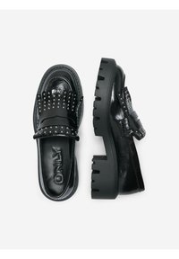Des chaussures noires en cuir style slip-on avec des clous sur le devant, une semelle épaisse et une finition lisse et brillante. Logo visible à l'intérieur.