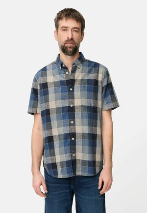 Mężczyzna z krótkimi brązowymi włosami i brodą, ubrany w niebiesko-szaro-beżową kratowaną koszulę na krótki rękaw z guzikami oraz niebieskie dżinsy na białym tle.