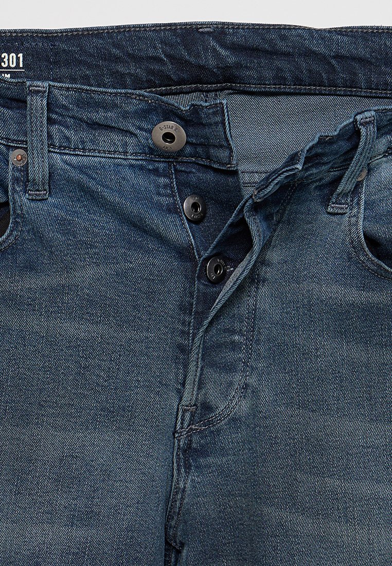 Jeans en denim bleu foncé avec une texture lisse. Les caractéristiques incluent une fermeture à bouton, cinq poches et des coutures contrastées le long des coutures.