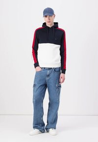 Navy, red, and white color-blocked hoodie with drawstrings, paired with blue baggy cargo jeans; white sneakers and a blue cap.