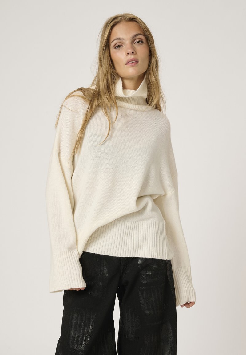 Cremefarbener, übergroßer Pullover mit hohem Kragen, geripptem Saum und Bündchen, aus einem weichen, strukturierten Stoff. Getragen mit dunklen gemusterten Hosen.
