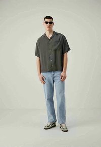 Grijze kortefe shirt met knopen, lichtblauwe jeans en groengetinte sneakers, gedragen door een model met een zonnebril tegen een neutrale achtergrond.