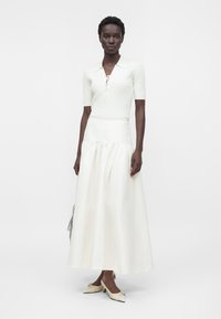 Femme portant un haut en maille blanc à manches courtes avec un col à lacets et une longue jupe blanche fluide, debout devant un fond uni.