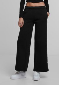 Črne široke trenirke z mehkim materialom, elastičnim pasom in stranskimi žepi. V kombinaciji z belimi supergami za sproščen videz.