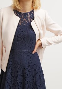 Light pink zip-up jacket over a navy blue lace dress, featuring a round neckline and floral lace pattern, with smooth fabric and pockets.