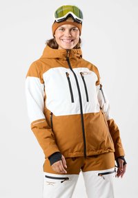 Giacca da sci arancione e bianca con cappuccio, dotata di due tasche con zip, una zip nera e dettagli con logo. Indossata con pantaloni da sci abbinati e occhiali da sci.