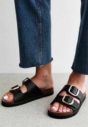 Czarne klapki na klamrze z korkową wkładką, płaską podeszwą i podwójnymi paskami. Noszone z niebieskimi dżinsami z postrzępionymi nogawkami na teksturowanej powierzchni.