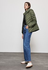 Veste matelassée vert olive avec capuche, fermeture éclair et poches latérales, associée à un jean large bleu et des baskets blanches sur un sol gris.