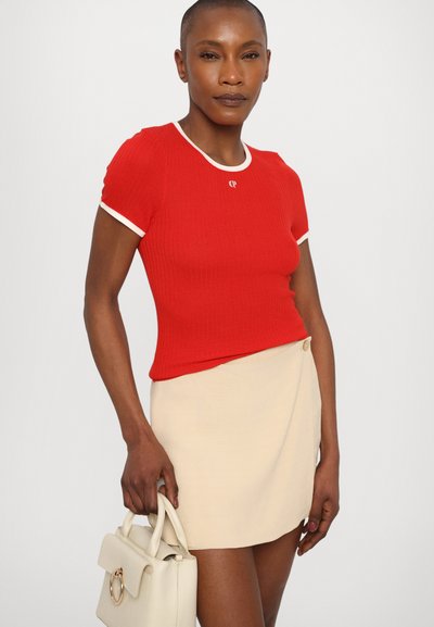 Haut rouge à côtes avec des manches courtes et un col blanc, associé à une mini-jupe beige, et portant un sac à main crème avec un accent rond doré.