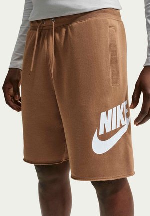 Brązowe spodenki Nike z białym logo, noszone przez osobę o ciemnej skórze i jasnej koszulce z długim rękawem, ręce luźno przy bokach.