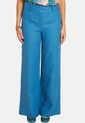 Woman wearing high-waisted, wide-leg blue pants with double buttons, paired with blue shoes and a colorful floral top partially visible.