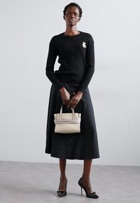 Pull noir à côtes, jupe noire à boutons et escarpins noirs. Tient un petit sac à main beige avec double poignées et un design horizontal.