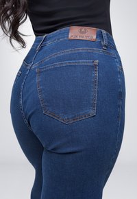 Donkerblauwe denim jeans met een hoge taille, oranje stiksels en een bruine leren patch met "FOX FACTOR" op de achterkant van de tailleband.