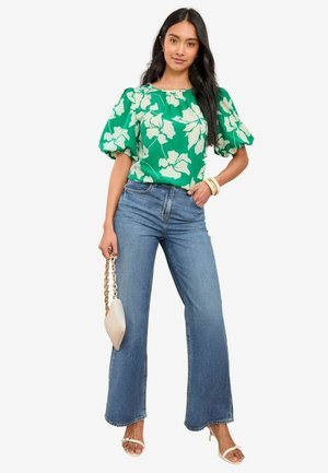 Grüne Bluse mit Blumenmuster und Puffärmeln, kombiniert mit hoch taillierten blauen, weit geschnittenen Jeans. Dazu eine kleine weiße Tasche und hellfarbige Sandalen.