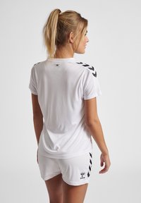 White athletic t-shirt with short sleeves, featuring black chevron stripes on shoulders and a logo on the shorts, made of breathable fabric.