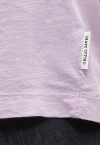 Lys lilla stoff med sydd kant og en hvit merkelapp merket "Marc O'Polo" synlig på sidesømmen over mørkt denimbånd.