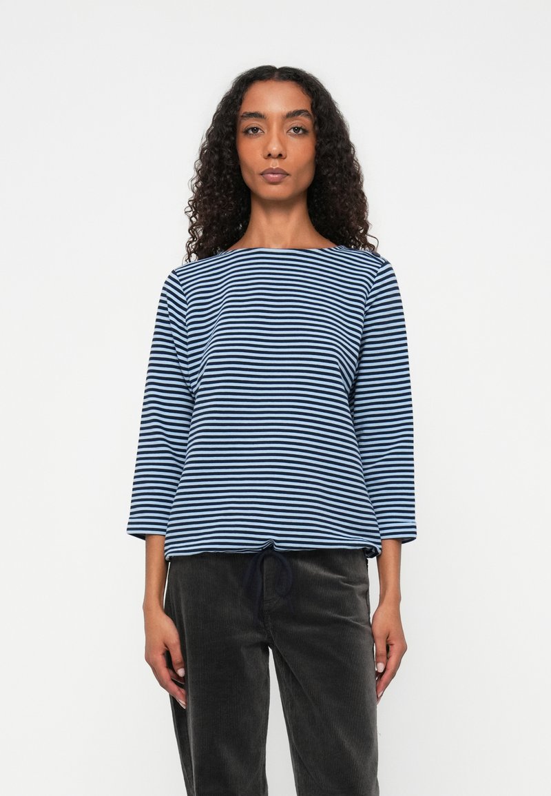 Blue and white striped long-sleeve top with a boat neckline, relaxed fit, and a drawstring detail at the hem, paired with gray corduroy pants.