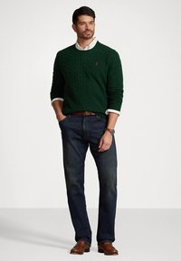 Πράσινο πουλόβερ με πλέξη καλτσοδέτας πάνω από ένα λευκό πουκάμισο, συνδυασμένο με σκούρα μπλε τζιν και καφέ δερμάτινα παπούτσια, με ένα απλό ρολόι.