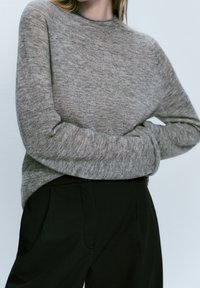 Harmaa neulotumpu, jossa on pyöreä kaula-aukko ja pitkät hihat, pehmeällä tekstuurilla. Yhdistetty mustiin räätälöityihin housuihin.