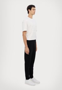 Weißes T-Shirt mit kurzen Ärmeln und Logodetail, kombiniert mit schwarzen Jogginghosen und weißen Turnschuhen, vor einem neutralen Hintergrund stehend.