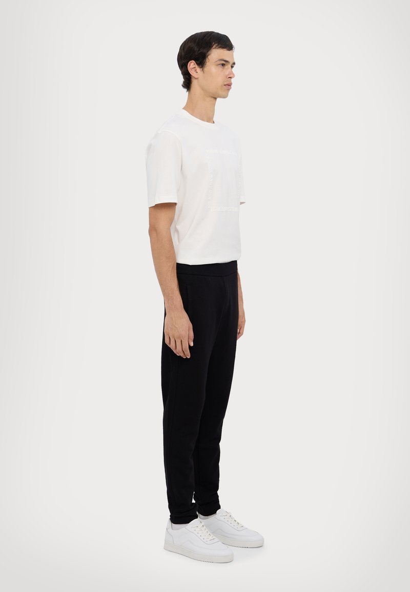Weißes T-Shirt mit kurzen Ärmeln und Logodetail, kombiniert mit schwarzen Jogginghosen und weißen Turnschuhen, vor einem neutralen Hintergrund stehend.