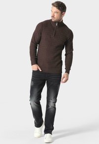 Brązowy dziany sweter z wysokim kołnierzem i zapięciem na zamek, w połączeniu z wyblakłymi czarnymi dżinsami i białymi sneakersami. Sweter ma teksturowany wzór.