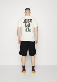 Camiseta de algodón blanca con un gráfico multicolor en la parte posterior que dice "¡SÓLO HAZLO!" y presenta varios diseños divertidos. Shorts negros y zapatillas naranjas.