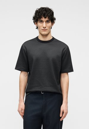 Άνδρας με σγουρά μαλλιά που φοράει μαύρο υφασμάτινο κοντομάνικο πουκάμισο και σκούρο παντελόνι με ρίγες, μπροστά σε απλό φωτεινό φόντο.