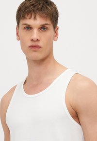 White cotton tank top with a ribbed texture and wide shoulder straps, featuring a round neckline and a close-fitting design.