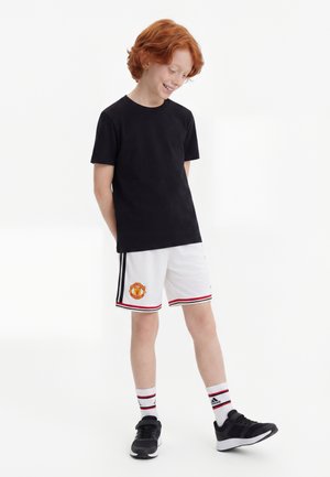 Gutt med rødt hår iført en svart t-skjorte, hvite Manchester United-shorts, hvite Adidas-sokker og svarte joggesko, stående mot en hvit bakgrunn.