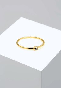Goldfarbene Metallring mit einem glatten Band und einem kleinen schwarzen Steinakzent auf einer Seite, auf einem weißen Präsentationssockel gesetzt.