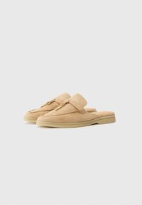 Suede beige slip-on shoes with a rounded toe, front strap, gold hardware accent, and a light-colored rubber sole. Flat and casual design.