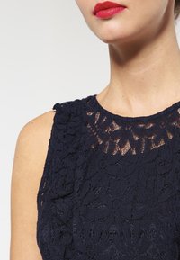 Navy blue lace dress featuring a floral pattern, sleeveless design, and a delicate, sheer neckline. Texture is intricate and detailed.