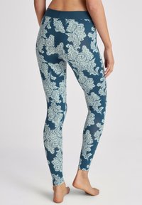 Femme portant des leggings bleus avec un motif floral paisley blanc, debout pieds nus, vue de dos sur fond blanc uni.