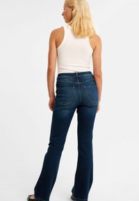 Dunkelblaue Schlagjeans mit hoher Taille, Denim-Material, Fünf-Taschen-Design und dezent kontrastierendem Nähen; Modell trägt ein weißes Tanktop.