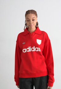 Felpa rossa con colletto, con il logo bianco "adidas" e un emblema triangolare. Realizzata in tessuto morbido, con maniche lunghe e una vestibilità comoda.