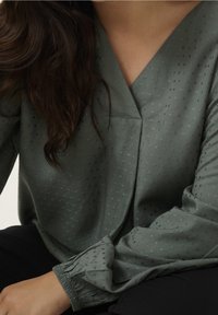 Blouse verte en tissu léger avec un décolleté en V, présentant un motif de points texturés et des manches longues froncées. Un pantalon noir est partiellement visible.