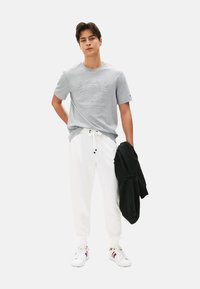 Camiseta de algodón gris con logo en relieve, pantalones jogger blancos con cordón, sudadera negra colgada del brazo y zapatillas blancas con acentos de color.