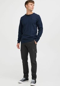 Granatowy sweter w serek wykonany z mieszanki bawełny; połączony z czarnymi spodniami cargo z bocznymi kieszeniami; noszony z czarnymi sneakersami i białymi sznurówkami.